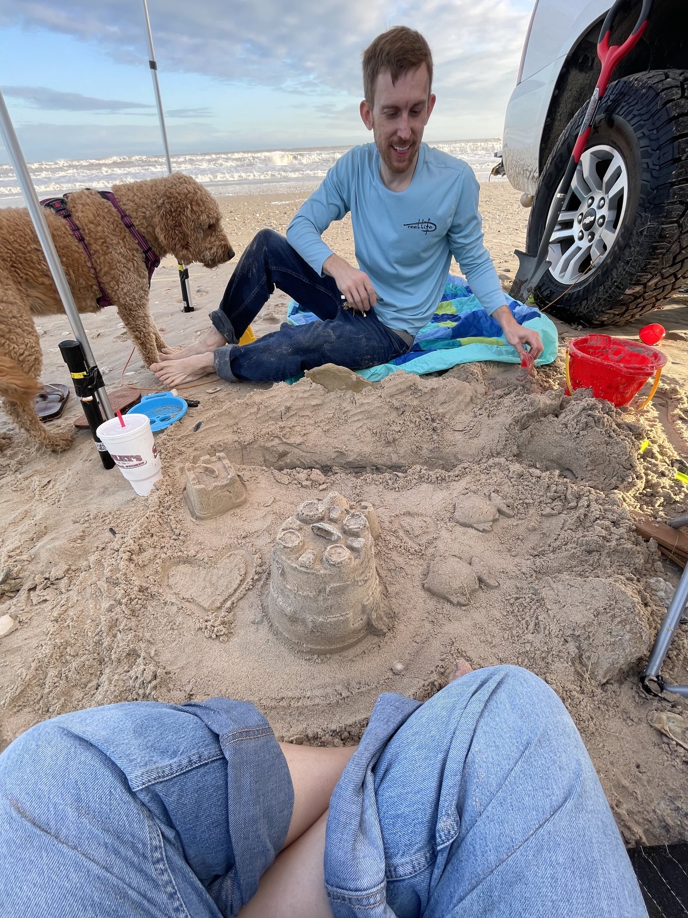 Jordan Polasek building a sandcastle on the beach with Autumn resting in the shade of the tent behind him, the ocean beyond